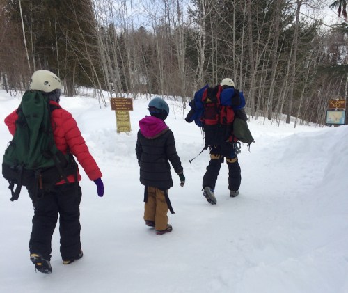 Three people in winter gear walking on a snowy trail, with trees and signs in the background.