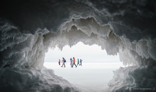 A group of people walks on a snowy landscape, framed by an icy cave entrance.