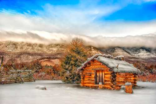 A cozy log cabin covered in snow, surrounded by a winter landscape and mountains under a blue sky with clouds.