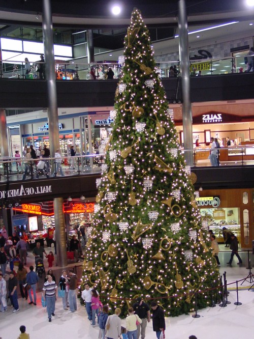 A large, decorated Christmas tree stands in a busy shopping mall, surrounded by people admiring it.