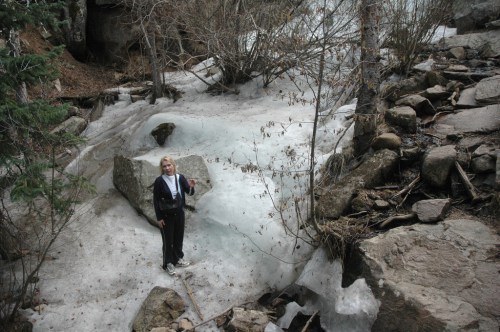 A person stands on rocky terrain near melting snow and ice, surrounded by trees and a natural landscape.