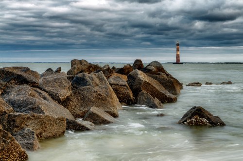 A rocky shoreline with waves gently lapping, featuring a distant lighthouse under a cloudy sky.