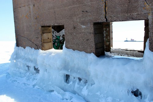 An abandoned structure partially covered in ice, with a view of a ship in the distance on a snowy landscape.