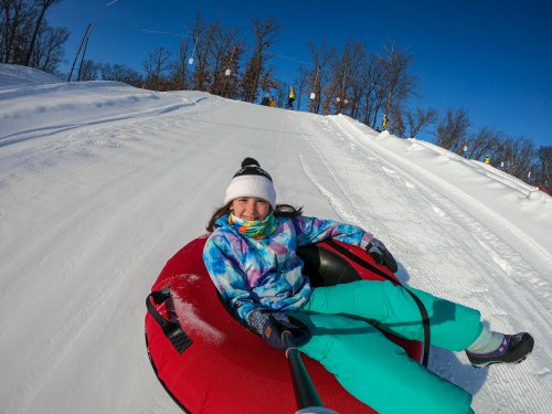 A girl in a colorful outfit smiles while tubing down a snowy slope on a sunny day.