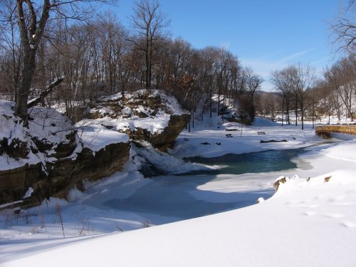 A snowy landscape featuring a river surrounded by rocky cliffs and bare trees under a clear blue sky.