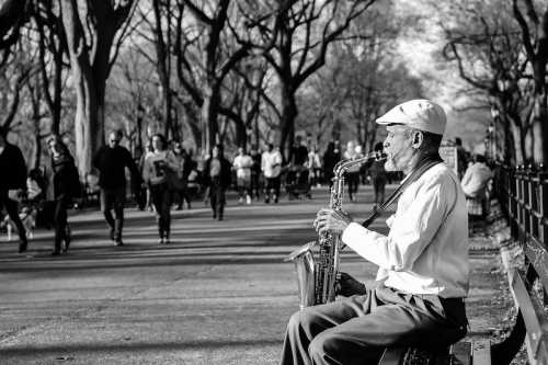 A saxophonist plays in a park as people walk by, surrounded by trees in a black and white scene.