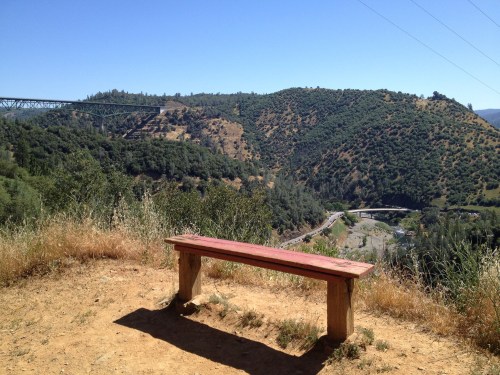 A wooden bench overlooks a scenic valley with hills and a bridge in the distance under a clear blue sky.