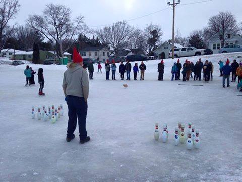 A group of people in winter attire play bowling on ice, with pins set up and a player preparing to roll a ball.