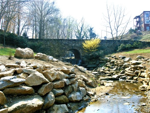 A stone bridge arches over a dry creek bed, surrounded by rocks and sparse trees in a sunny park setting.