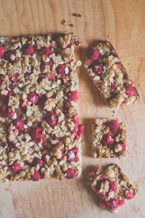 A wooden surface with a rectangular oat bar topped with raspberries, with a few pieces cut and scattered nearby.