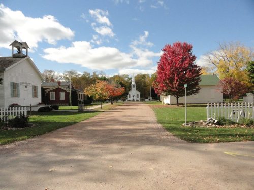 A peaceful rural scene with a gravel road, colorful trees, and a white church in the background under a blue sky.
