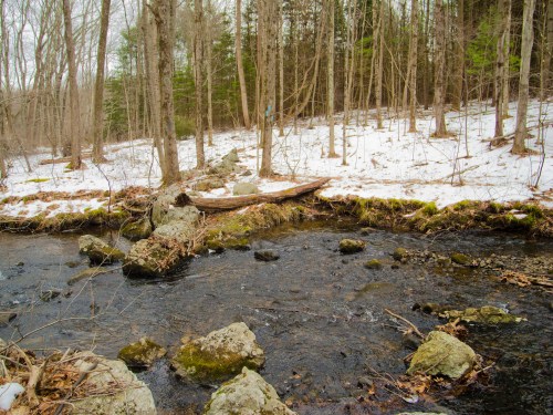 A serene winter scene with a flowing stream, rocky banks, and bare trees in a snowy forest.