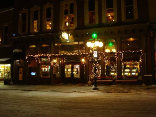 A cozy storefront decorated with lights and a Christmas tree, set in a snowy street at night.