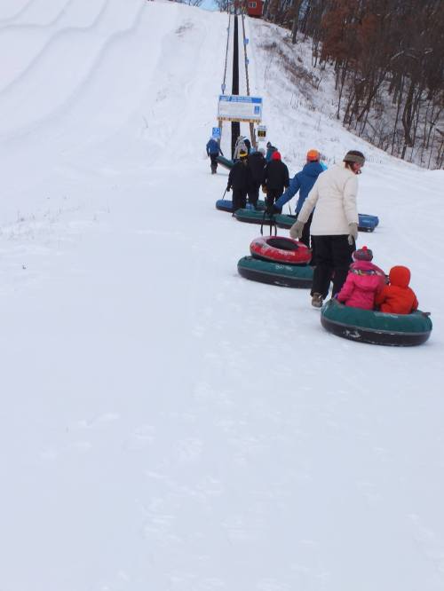 A group of people in winter clothing are walking up a snowy hill, pulling inflatable tubes behind them.