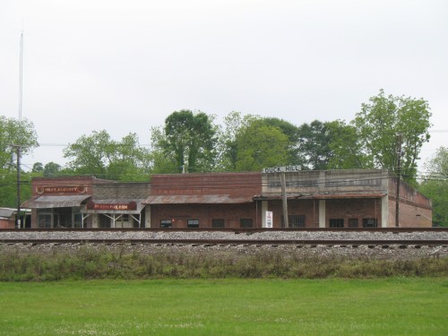 Old brick buildings along a railway track, surrounded by greenery and a cloudy sky.