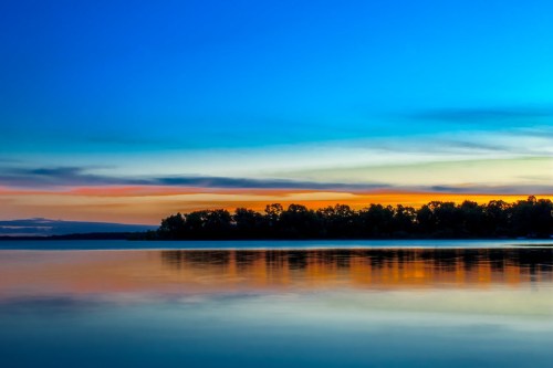 A serene lake at sunset, reflecting vibrant blue and orange hues in the sky and water, surrounded by trees.