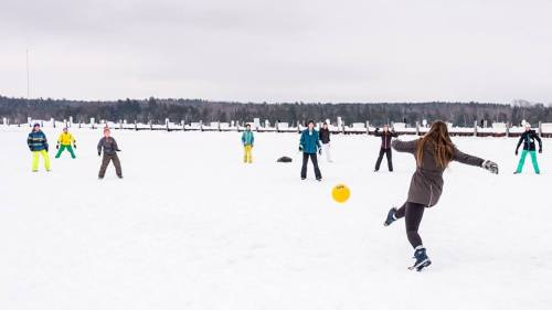 A group of people playing a game on a snowy field, with one person kicking a yellow ball while others watch.