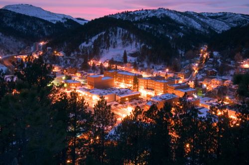 A scenic view of a snowy town at dusk, illuminated by warm lights, surrounded by mountains and trees.