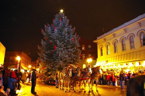 A festive scene with a large Christmas tree, horse-drawn carriage, and crowds enjoying holiday decorations at night.