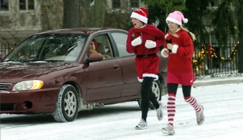 Two people in festive outfits run on a snowy street, while a car drives by in the background.