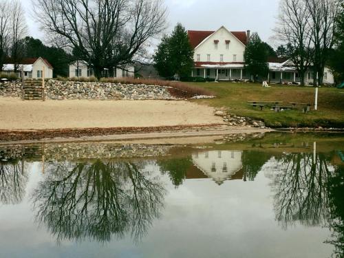 A serene lakeside view with a sandy beach, trees, and a house reflecting in the calm water.