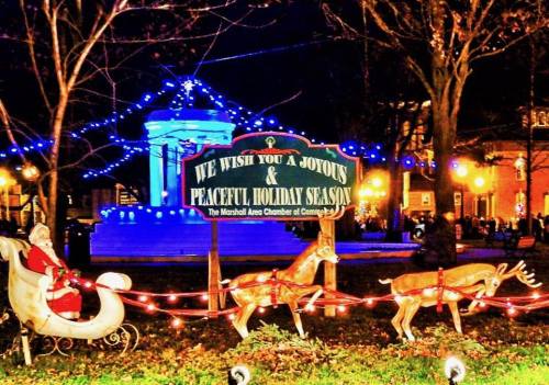 A festive holiday sign with Santa in a sleigh and reindeer, surrounded by colorful lights in a park setting.