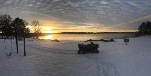 A person rides an ATV on snowy ground near a lake at sunset, with trees and a swing set in the background.