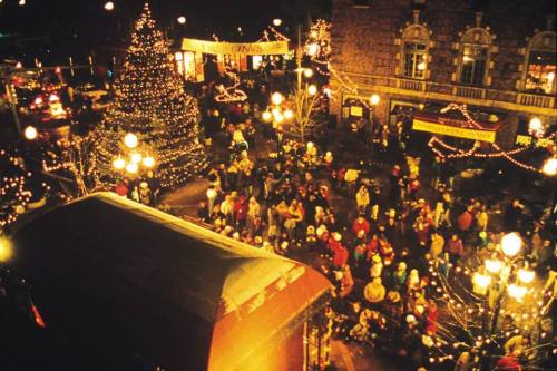 A festive night scene with a large Christmas tree, crowds of people, and colorful lights illuminating the area.