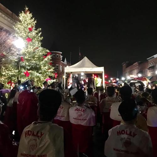 A marching band performs under festive lights near a decorated Christmas tree during a nighttime event.