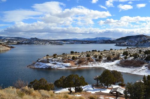A serene lake surrounded by snow-dusted hills and greenery under a partly cloudy sky.