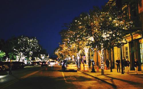 A festive street at night, lined with trees adorned with twinkling lights and bustling with people and cars.