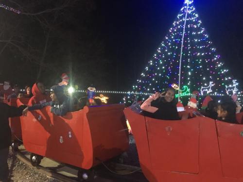 A festive scene with children in red train cars near a brightly lit Christmas tree at night.
