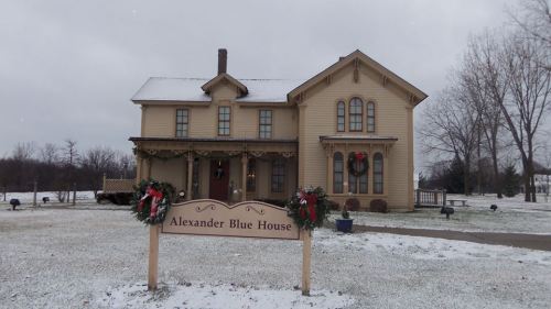 A two-story yellow house with holiday decorations and a sign reading "Alexander Blue House" in a snowy landscape.