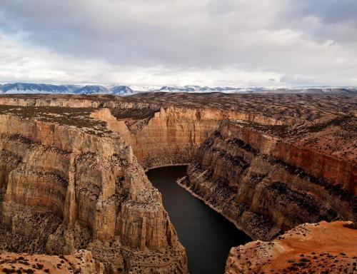 A winding river flows through a deep canyon, surrounded by rugged cliffs and distant snow-capped mountains under a cloudy sky.