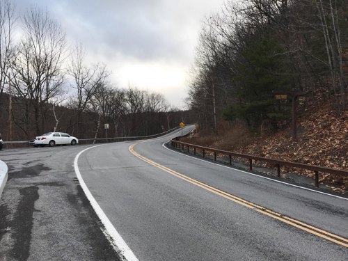 A winding road curves through a wooded area, with a parking area visible on the left and cloudy skies above.