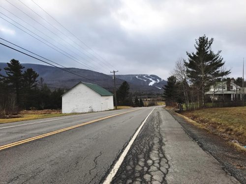 A quiet road lined with trees, leading to a distant mountain with ski trails, under a cloudy sky.