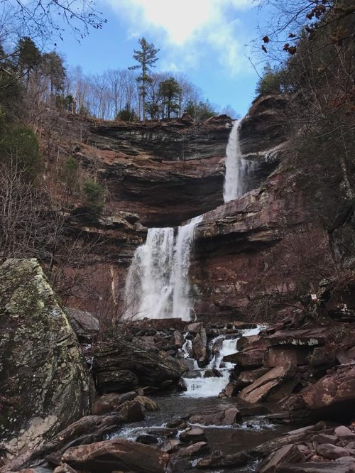 A scenic waterfall cascading down rocky cliffs, surrounded by trees and a blue sky.