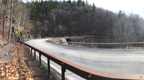 A winding road curves through a forested area, with bare trees and a guardrail along the edge.