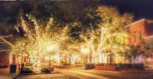 A charming street illuminated by festive lights on trees and buildings, creating a warm, inviting nighttime atmosphere.