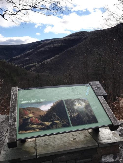 A scenic view of Kaaterskill Clove with a sign displaying artwork and information about the area, surrounded by mountains.