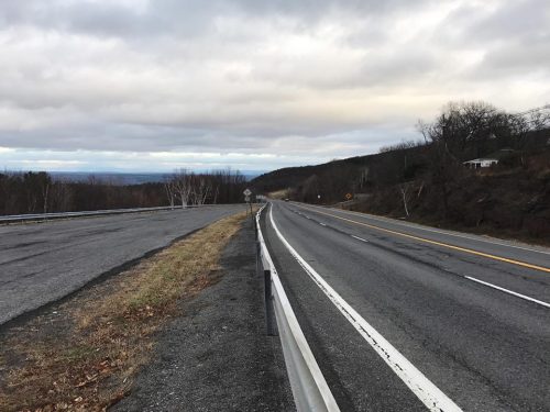 A wide, empty road stretches into the distance, flanked by grassy areas and hills under a cloudy sky.