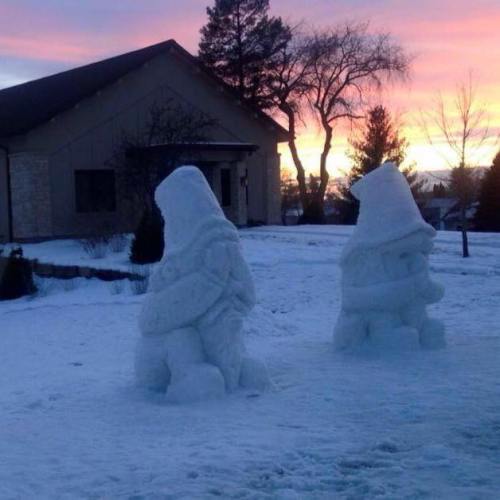 Two snow sculptures resembling figures stand in a snowy yard, with a colorful sunset in the background.