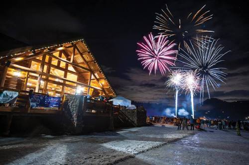 A cozy wooden cabin illuminated at night, with colorful fireworks bursting in the sky above.