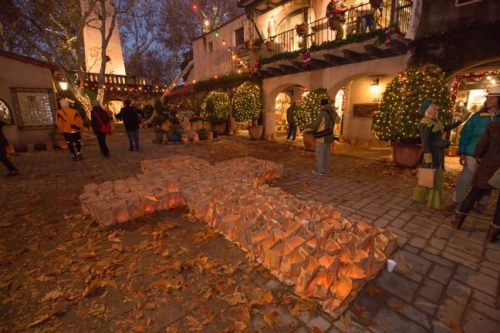 A festive outdoor scene with holiday lights, decorated trees, and people walking around a large illuminated cross made of bags.