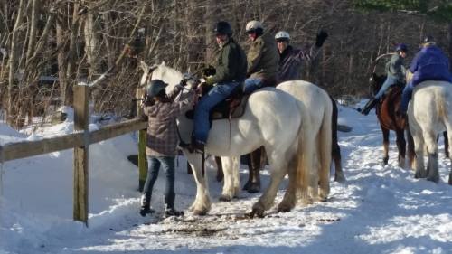 A group of people on horseback in a snowy landscape, with one person assisting another on a white horse.