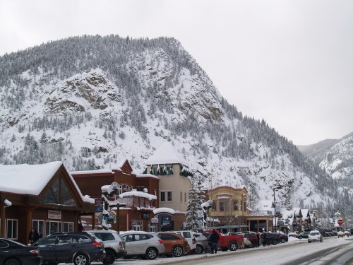 Snow-covered mountain backdrop with a quaint town street lined with shops and parked cars.