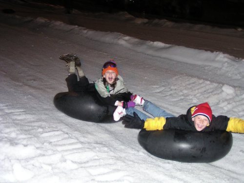 Two children joyfully tubing down a snowy slope, wearing winter gear and smiling in the night.
