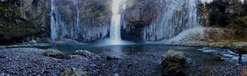 A panoramic view of a waterfall cascading into a rocky pool, surrounded by lush greenery and ice formations.