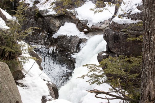 A frozen waterfall cascading over rocky terrain, surrounded by snow and evergreen trees.