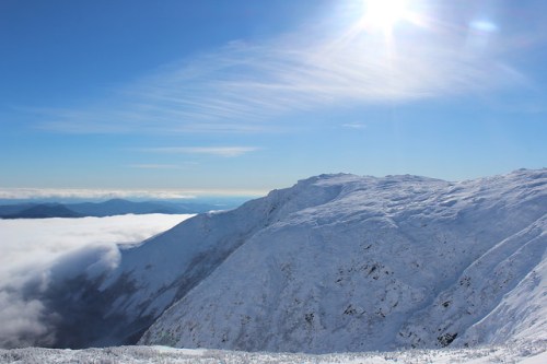 A snowy mountain landscape under a bright sun, with clouds rolling in the valley below.